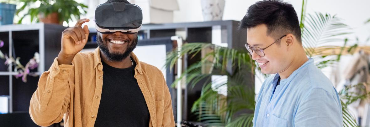 Colleagues testing out virtual reality device inside the metaverse - stock photo  African man wearing virtual reality glasses and gesturing with ale colleague standing by using digital tablet in office. Men testing out virtual reality device at office.