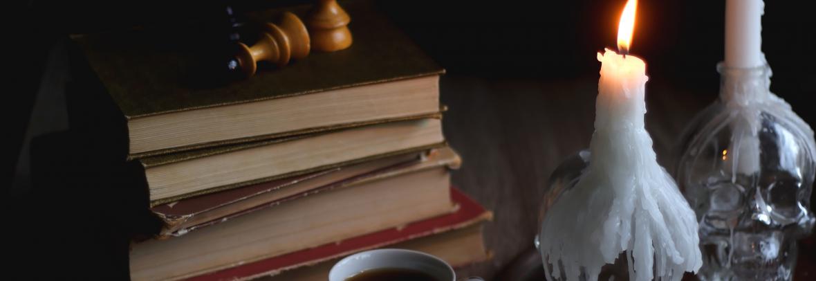 Stack of vintage books, cup of tea or coffee, lit candles, reading glasses and chess pieces on wooden table. Dark academia concept. Selective focus.