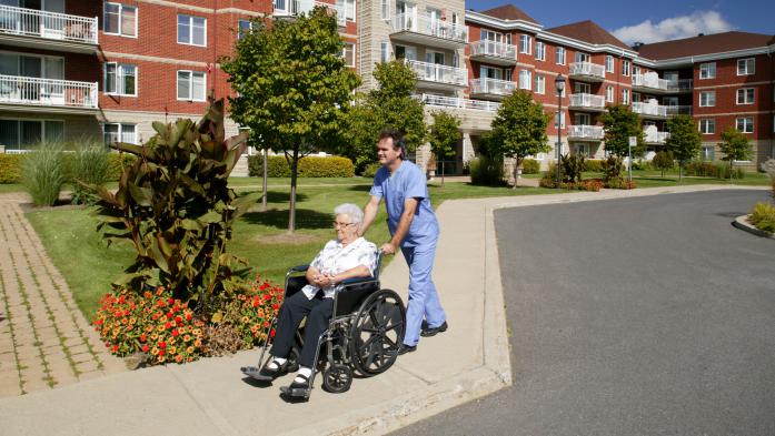 Nurse pushes an older woman in a wheelchair in front of a senior living home