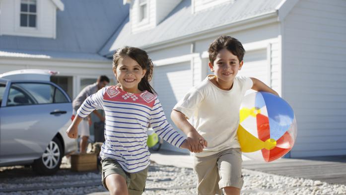 Brother and sister kids with beach ball running toward beach with house in background