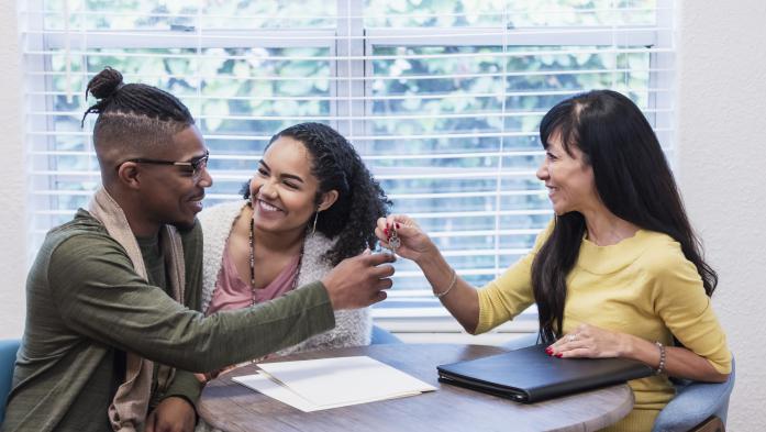 Young couple meeting with Real Estate Agent, handing over key - stock photo  A young African-American couple meeting with a real estate agent, a mature mixed race Chinese and Caucasian woman, sitting at a table by a window. They are smiling at each other and the Real Estate Agent is handing over the key to their new home or rental.