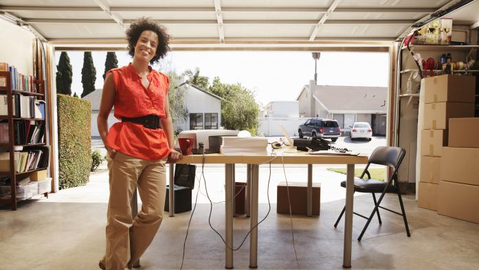 Woman in office space in garage, portrait. Woman in orange shirt and tan pants standing near a desk in her garage.