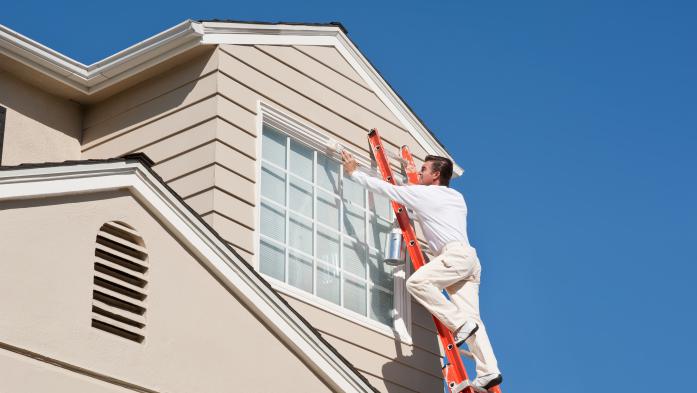 House painter with paint brush painting the trim of a 2nd story window of a Cape Cod Style house.