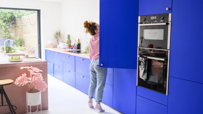 Full length view of woman in jeans and casual top standing on tiptoe to access high shelf in vibrant blue kitchen.